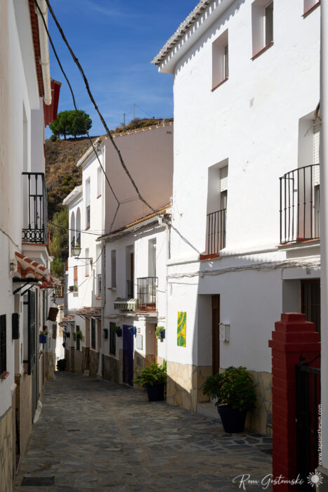 A narrow cobblestone street in &Aacute;rchez is lined with white buildings, colourful artwork, balconies, and potted plants. Overhead wires cross the scene, with a hill of greenery visible in the background beneath a clear blue sky.