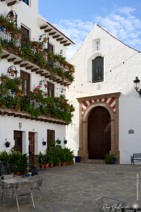 A charming courtyard with a whitewashed building covered in flower-filled balconies stands next to the Iglesia de Nuestra Se&ntilde;ora de la Expectaci&oacute;n, its large wooden door and arched entrance framed by a blue sky with scattered clouds.