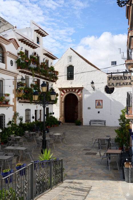 The square in Canillas de Albaida is a quiet spot with empty tables and chairs, surrounded by white buildings with balconies and plants. A church with an arched doorway stands in the background under a partly cloudy sky.