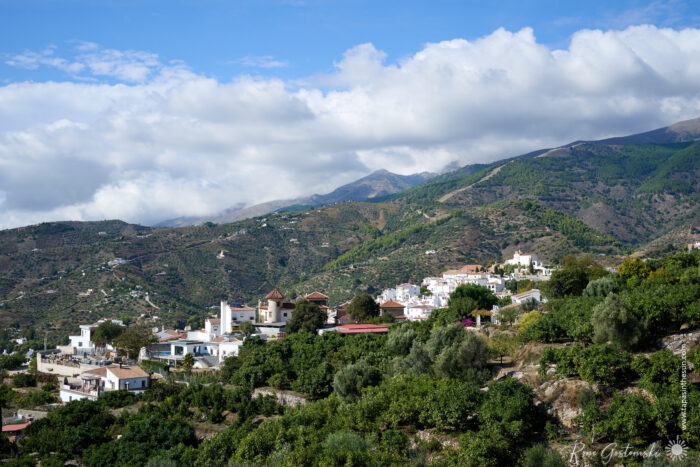 A hillside village, Canillas de Albaida, with white houses surrounded by green trees and mountains under a partly cloudy sky.