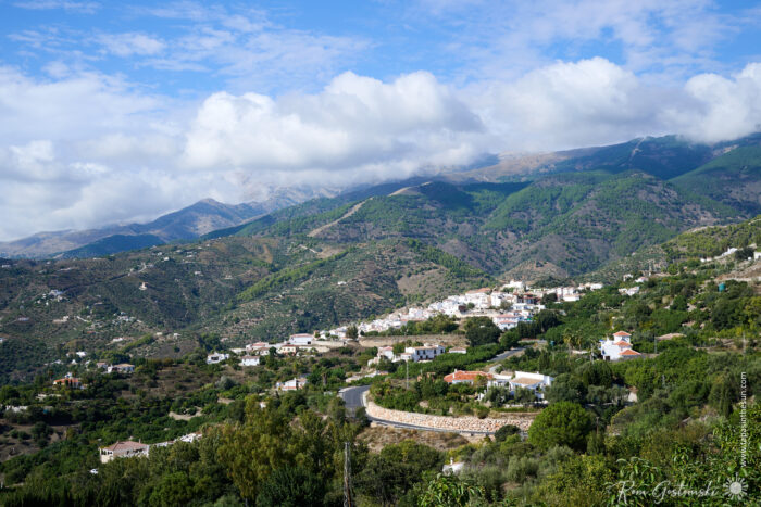 A scenic view of Canillas de Albaida nestled in green hills with winding roads, white houses, and mountains in the background under a partly cloudy blue sky.