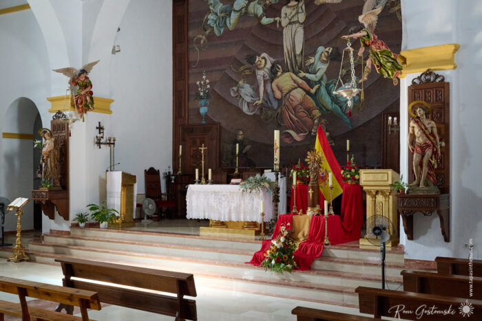 The interior of a church features wooden pews and the altar inside Parroquia Nuestra Se&ntilde;ora de la Asunci&oacute;n, adorned with red and gold cloth, floral arrangements, religious statues, and a mural of angels behind the ornate altar.