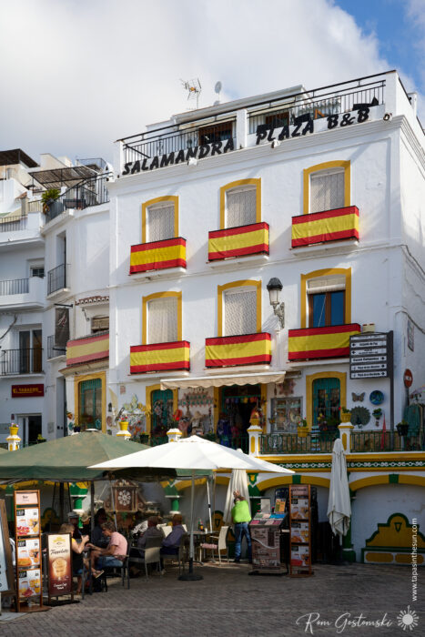 A white building with yellow window frames and red-and-yellow Spanish flags, labelled Salamandra Plaza B&amp;B, sits in the main square in C&oacute;mpeta. People relax at tables outside under umbrellas in the lively plaza with blue sky above.