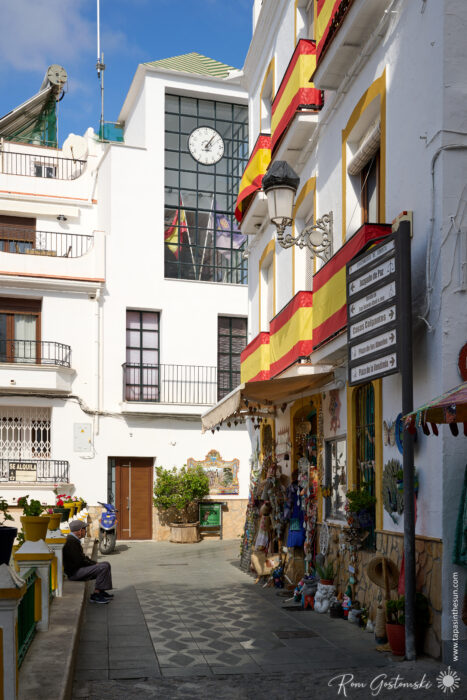 A narrow alley along one side of the main square in C&oacute;mpeta with white buildings draped in Spanish flags, a shop offering souvenirs, and a few people sitting and walking. In the background stands a modern glass building with a clock.
