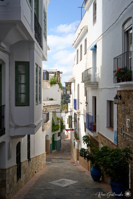 A narrow cobblestone street in C&oacute;mpeta lined with white buildings, black railings, small balconies, and potted plants stretches under a partly cloudy sky in this charming Mediterranean town.
