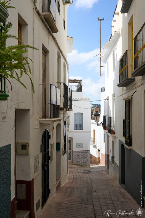 A narrow cobblestone street in C&oacute;mpeta lined with white buildings, black railings, small balconies, and potted plants stretches under a partly cloudy sky in this charming Mediterranean town.