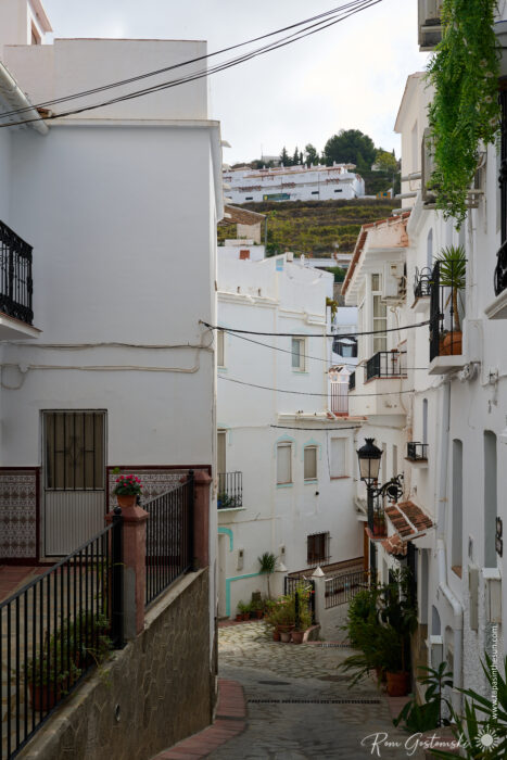 Narrow, winding street lined with whitewashed buildings, potted plants, and balconies in the Mediterranean village of C&oacute;mpeta; distant white houses and greenery dot a hillside beneath a cloudy sky.