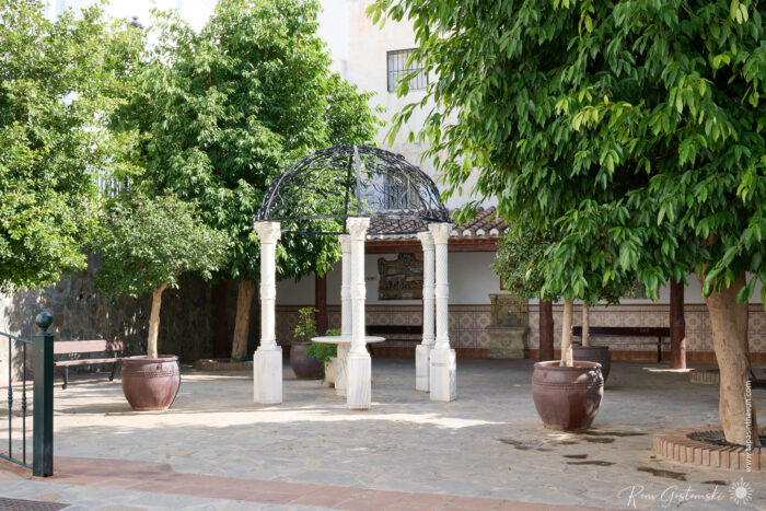 A sunlit courtyard in C&oacute;mpeta, with a decorative wrought-iron gazebo supported by stone columns, surrounded by large potted trees, tiled benches, and a mosaic wall in the background.
