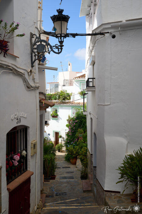 A narrow, sunlit alleyway in C&oacute;mpeta, a Mediterranean village lined with whitewashed buildings, potted plants, a black iron lantern, and balconies. The path leads down towards more houses under a bright blue sky.