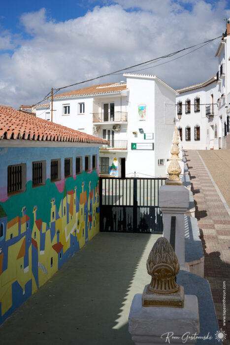 A vibrant mural of colourful houses decorates the school wall in sunny C&oacute;mpeta, featuring white buildings, balconies, and a clear blue sky with scattered clouds overhead.
