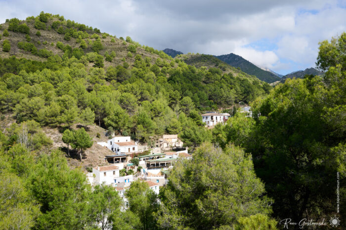 A small cluster of white houses in El Acebuchal nestles among green, tree-covered hills under a partly cloudy sky. The buildings are surrounded by dense forest and mountains in the background.