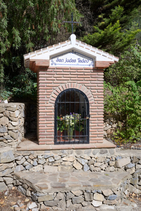 Small brick shrine with a tiled roof and a sign reading St Jadas Tadeo. The shrine has a black metal gate and contains flowers inside. It is surrounded by stone walls and greenery.