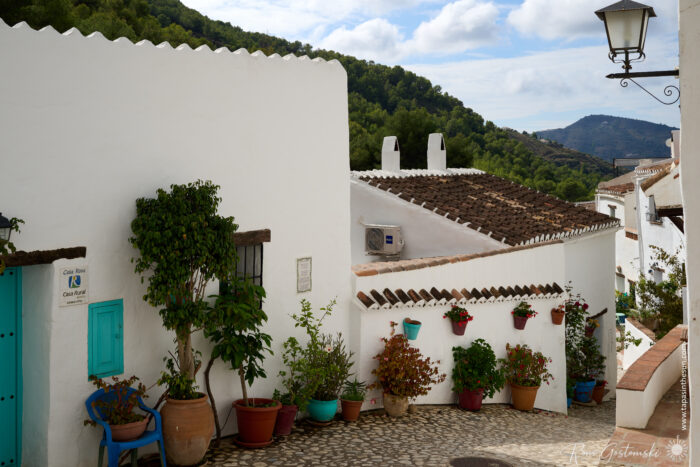 A narrow cobbled street in El Acebuchal&mdash;known as the lost village&mdash;winds between whitewashed houses, potted plants, and a traditional streetlamp, all set against green hills and a partly cloudy sky.