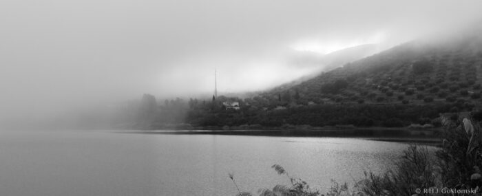 A black-and-white photo of a misty lakeshore with hills in the background, partially obscured by fog. A faint outline of trees, a house, and a tall aerial are visible through the mist across the still water.