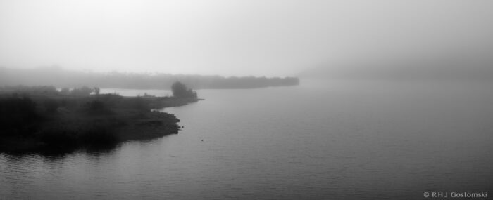 A calm lake with a foggy shoreline, where trees and landscape fade into the mist, creating a serene and tranquil black-and-white scene.