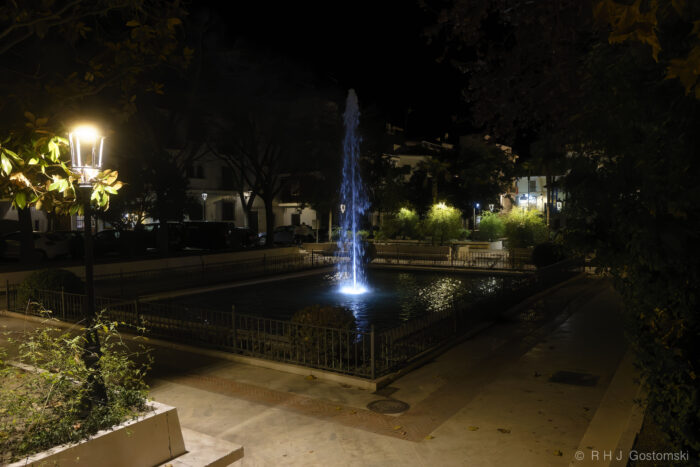 Plaza Puerta del Sol A square fountain illuminated by blue lights at night sits in Plaza Puerta del Sol, surrounded by trees and benches. Street lamps cast a warm glow on the walkway, creating a calm and quiet atmosphere.