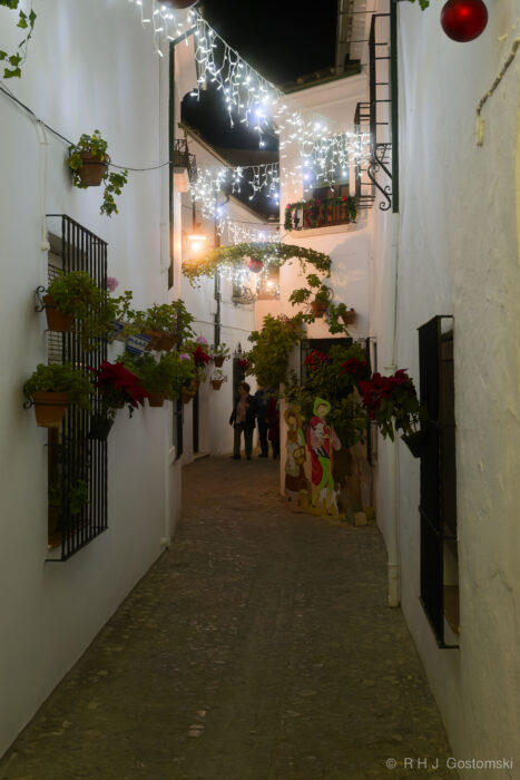 A narrow, cobblestone alley in Barrio de La Villa A narrow, cobblestone alley in Barrio de La Villa, Priego de Córdoba with white walls is decorated with Christmas decorations—hanging plants, poinsettias, festive lights, and a nativity scene—as a few people gather in the background at night.