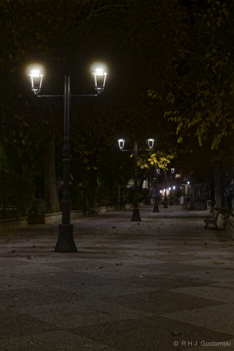 A pedestrianised walkway in Priego de Córdoba at night A quiet, empty park pathway at night in Priego de Córdoba, lined with tall, glowing streetlamps and trees with autumn leaves. Benches are visible along the path, and soft light creates a peaceful atmosphere.