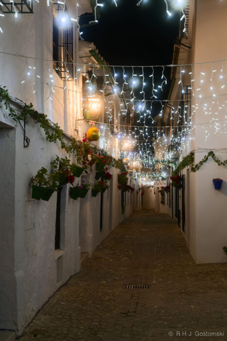 Barrio de La Villa, Priego de Córdoba, Christmas decorations A narrow cobbled street in Priego de Córdoba, lined with white buildings adorned with hanging plants and sparkling Christmas decorations, glows softly under strings of white fairy lights at night.