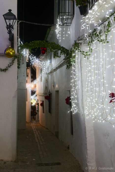 Barrio de La Villa district, Priego de Córdoba. Christmas lights in an alley A narrow, cobbled street in Priego de Córdoba with white walls is decorated with hanging Christmas lights, greenery, and festive ornaments at night, creating a jolly and cosy atmosphere.