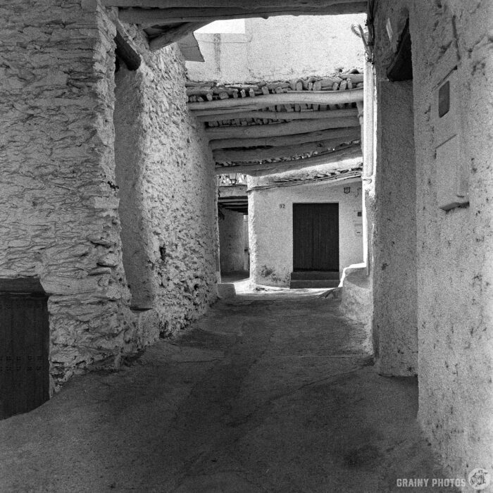 Narrow Alley Under a Series of Tinaos in Capilerilla A narrow, winding alleyway in Capilerilla, Spain, with white stone walls and a rustic wooden tinao, leads to a closed wooden door; shadows and light create a peaceful, timeless atmosphere in this black and white photo.