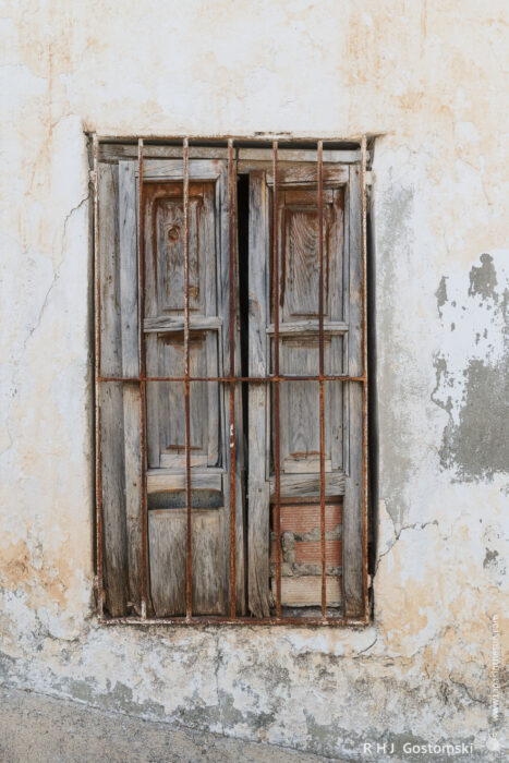 Shuttered window showing decay A weathered, wooden window with rusty metal bars set in a cracked, peeling plaster wall. The wood is aged and faded, adding a rustic and abandoned feel to the scene.