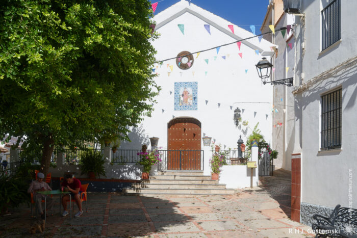 Iglesia San Lorenzo A small white church, Iglesia San Lorenzo, with a decorated entrance is adorned with flowers and colourful flags. Two people sit at a table in the shade of a large tree in the sunlit courtyard.