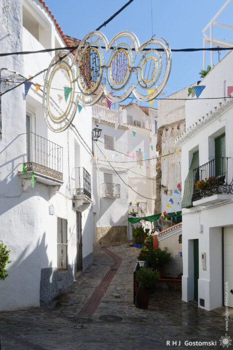 Fiesta decorations A narrow cobbled street winds through the whitewashed Spanish village of Guájar-Faragüit, decorated with colourful fiesta decorations and circular light fittings overhead, all bathed in bright sunlight.