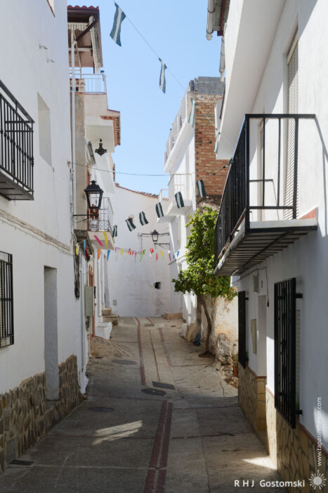 A narrow street and white houses in Guájar-Faragüit A narrow, sunlit street in Guájar-Faragüit with whitewashed buildings, black balconies, a tree, and festive pennant bunting hanging across the alleyway. Shadows and flags add charm to the peaceful scene.
