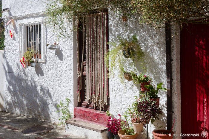 Front door A white stucco house with red accents, potted plants, and hanging greenery by the entrance, featuring a doorway with a beaded curtain and sunlight casting shadows on the wall.