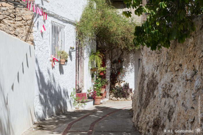Narrow, sunlit alley A narrow, sunlit alley with white and stone walls, potted plants, and small colourful flags hanging by a window, creating a charming and peaceful Spanish atmosphere.