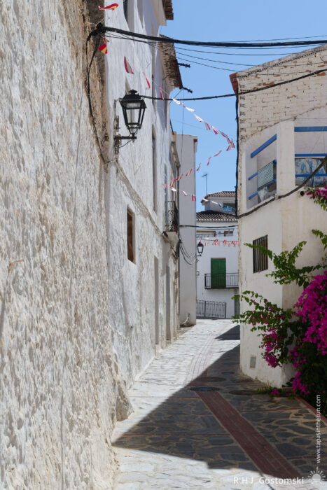 A narrow street and white houses in Guájar-Faragüit A narrow, sunlit alleyway with whitewashed walls, fairy lights, and flowering bougainvillaea on the right. The stone pathway curves, leading to a building with green shutters in the background.