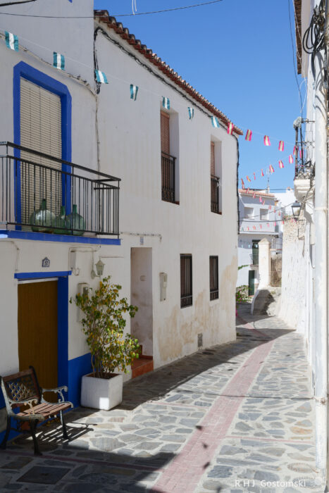 A narrow street and white houses in Guájar-Faragüit A narrow, sunlit alley with whitewashed buildings, blue trim, balconies, and festive bunting overhead. A bench and potted plants sit near a wooden door on the left side.