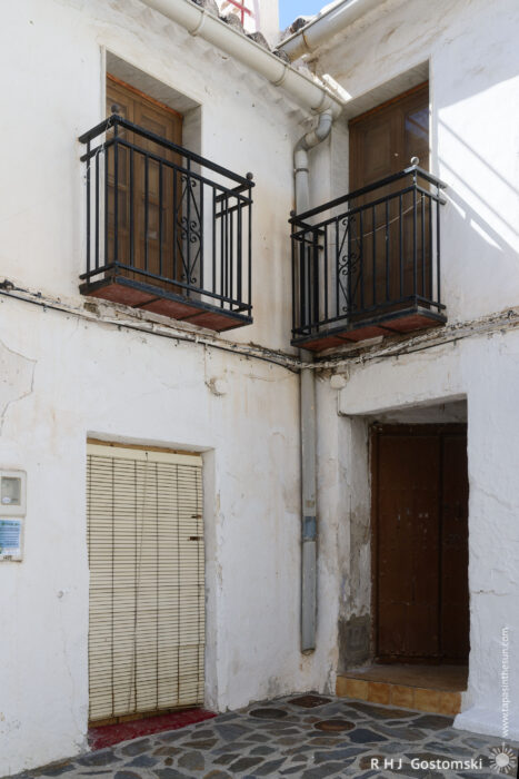 Village houses A white-walled building with two small balconies featuring black railings, both above a ground floor with a closed brown door and a window covered by beige blinds. A drainpipe runs between the balconies.