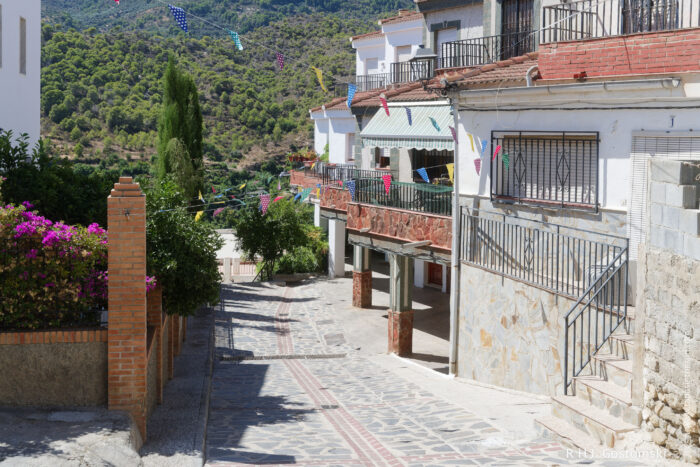 Guájar-Faragüit A quiet, sunlit street in a hillside village features stone pavement, brick and stone houses with balconies, colourful pennant banners, and lush greenery in the background.