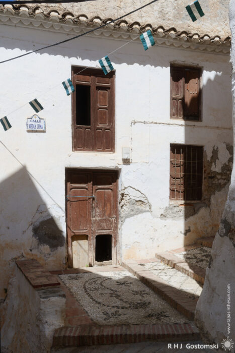 An abandoned house A rustic white abandoned house with weathered wooden doors and windows, mosaic stone steps, and green-and-white flags strung above. Shadows fall across the façade, and a street sign reads Calle Media Luna.