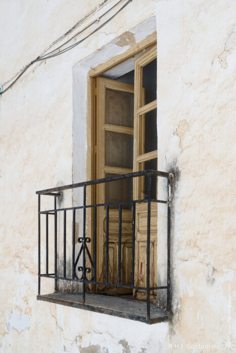 A Juliet balconny A weathered wooden door with peeling paint stands open behind a small wrought-iron balcony on a faded, textured white wall. Exposed wires run above the door, giving the scene a rustic appearance.