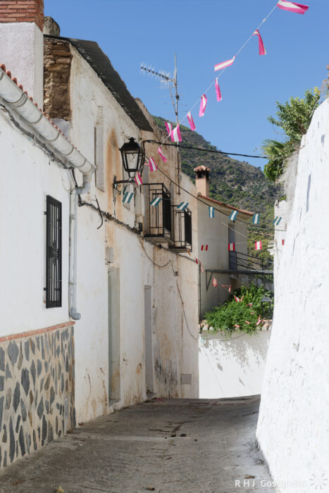 Guájar-Faragüit A narrow street in a Spanish village with whitewashed walls, colourful pennant flags strung above, an old lamp, and a view of distant hills under a clear blue sky.