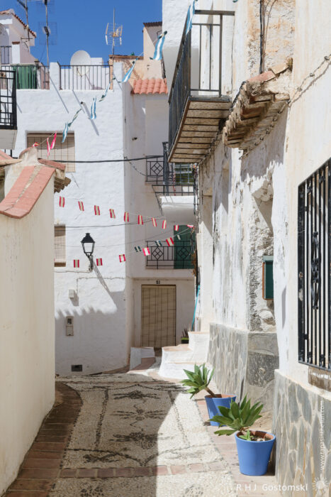 A narrow street in Guájar-Faragüit A narrow, sunlit alley in Guájar-Faragüit, a Spanish village with whitewashed walls, tiled balconies, potted plants, and colourful festive bunting strung across the street. The ground is decorated with a stone mosaic pattern.