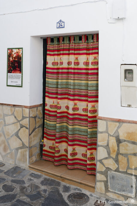 An ornamental fabric curtain over the front door A doorway with a colourful, patterned curtain hangs in place of a door under a white wall marked “34.” The lower wall is decorated with tan stonework, and there is a framed picture and plaque beside the entrance.