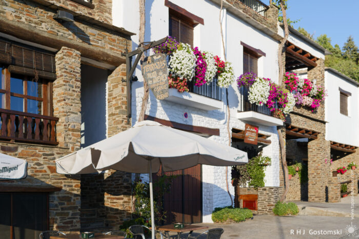 Flower boxes on balconies Sunlit street café in Capileira featuring a whitewashed and stone-built house with wooden balconies and iron railings overflowing with pink, purple and white hanging flowers, a large cream parasol shading outdoor tables and a rustic wooden sign above the doorway.