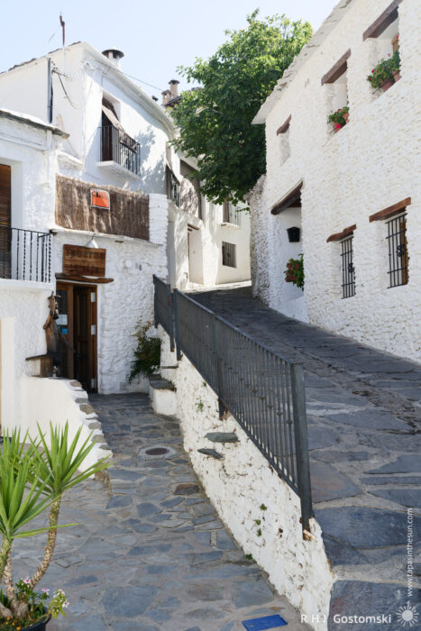 A narrow, sloping stone street between white houses A narrow, sloping stone street winds between whitewashed buildings with wooden balconies and colourful flower pots in the sunny Spanish village of Capileira.