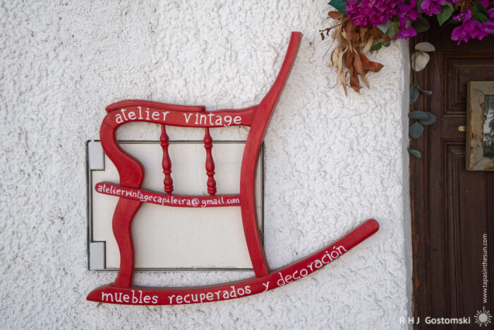 Artisan furniture restoration and art A red rocking chair frame is mounted on a white textured wall with text promoting atelier vintage, furniture restoration, and decoration services. Purple flowers hang above, partially visible near a wooden door.