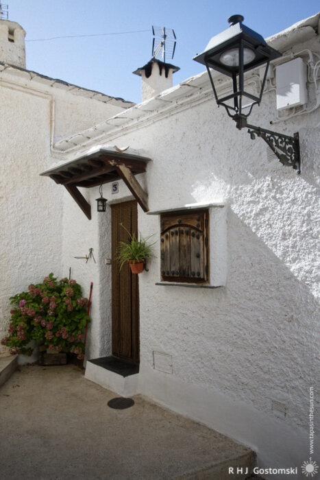 The front door to a small house A whitewashed house with a dark wooden door, a small shuttered window, a black metal lantern, and a hanging plant. Pink flowers in a pot sit on the ground near the entrance. Bright daylight illuminates the scene.