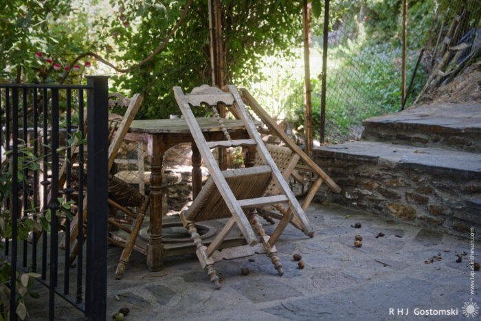 Table and chairs A rustic outdoor scene with a wooden table and four chairs leaning against it, next to a black metal fence and stone steps, surrounded by greenery and dappled sunlight. Walnuts lie on the ground.