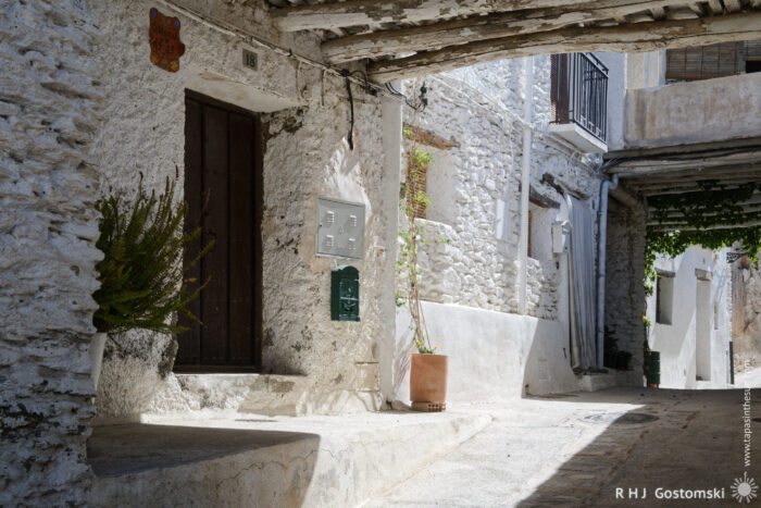 Tinaos over a narrow street A narrow, sunlit street with white stone walls and a wooden door, potted plants, and rustic wooden beams forming a traditional tinao overhead in Capileira.
