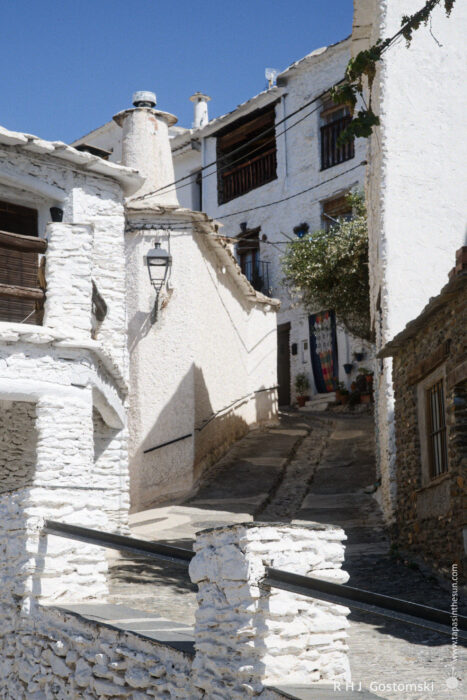 An alley with twists and turns A narrow, steep alley with whitewashed stone houses, wooden balconies, and a small tree under a clear blue sky in Capileira. A colourful curtain hangs over a door.
