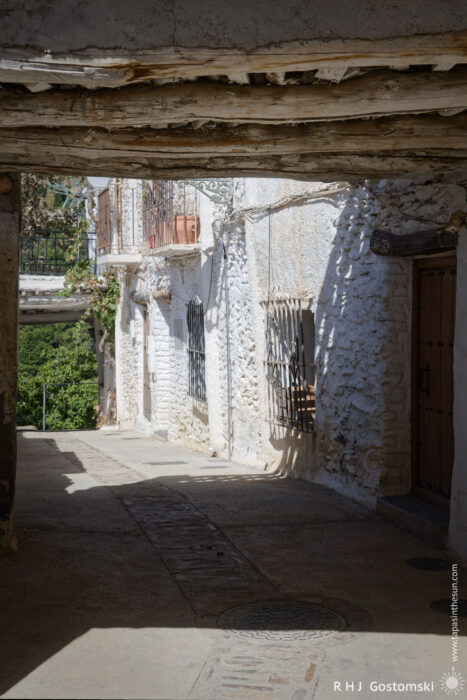 Tinaos over a narrow street A narrow street in Capileira, lined with whitewashed stone buildings, wooden-beamed tinaos overhead, windows with burglar bars, and sunlight casting shadows across the path.