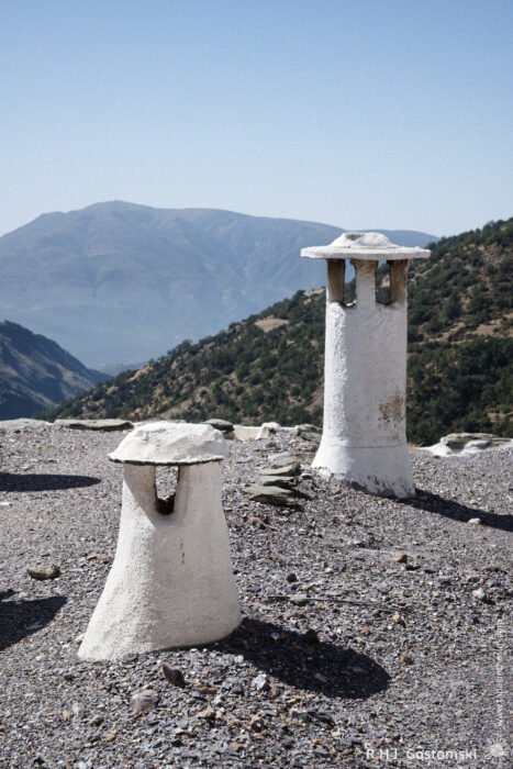 Alpujarran chimneys Two white, cylindrical Alpujarran chimneys with conical caps stand on a traditional flat rooftop overlooking forested mountains and a hazy blue sky in the background.