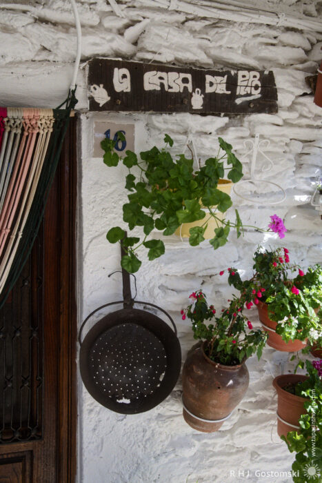 Potted plants and a rusted through colander A rustic white wall displays a wooden sign reading La casa del Pipo, with potted plants, a brown ceramic jug, a metal colander hanging below, and a striped curtain partially covering a wooden door.